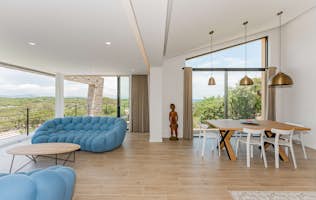 location - Thalassa Salins - Modern dining room with panoramic view  location - Thalassa Salins - This modern dining room offers a panoramic view of the green hills. It is furnished with a blue sofa, a wooden dining table, and white chairs, creating a warm and inviting atmosphere.