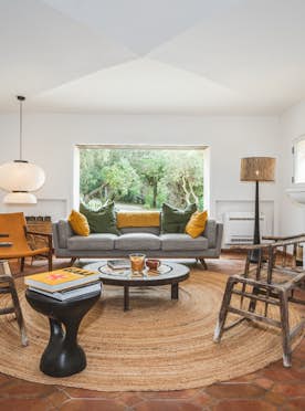 location - Barbarine - Modern living room with fireplace and large window  location - Barbarine - A modern and elegant living room with a fireplace and a large window offering a view of the garden. The furniture includes a grey sofa, wooden armchairs, and colorful cushions.