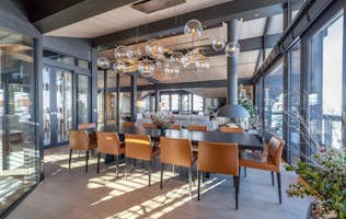 accommodation - Chalet B  - Modern dining room with designer furniture  accommodation - Chalet B  - This image shows a modern dining room with a wooden table surrounded by brown leather chairs. The room is lit by a designer chandelier and large windows offering a panoramic view.