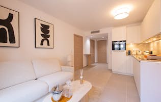 location - Oscar - Modern living room and open kitchen  location - Oscar - This image shows a modern living room with a white sofa and an open kitchen equipped with modern appliances. The space is bright and well-decorated with abstract artwork.