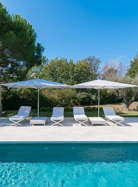 location - Amandiers - Pool with terrace and outdoor furniture  location - Amandiers - Image of an outdoor pool with a stone terrace, lounge chairs, and an umbrella, surrounded by lush greenery.