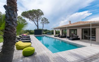 location - Salins - Modern pool and terrace with garden view  location - Salins - This image shows a modern pool surrounded by a wooden terrace with lounge chairs and colorful cushions, offering a stunning view of a lush garden and trees.