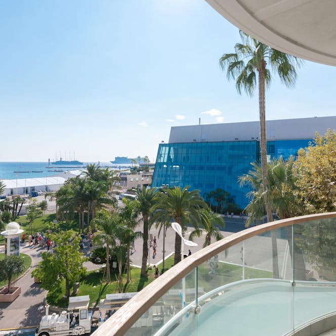 location - Croisette - Panoramic view from a balcony with sea view  location - Croisette - Image of a balcony offering a stunning view of the sea, palm trees, and a modern building. Ideal for a luxurious lifestyle.