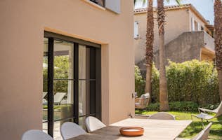 location - Noria - Modern outdoor dining terrace with wooden table  location - Noria - Image of a modern outdoor dining terrace with a wooden table and white chairs, surrounded by palm trees and lush greenery.