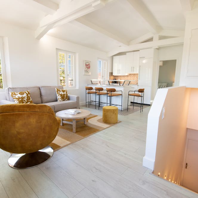 location - Riviera - Modern and Bright Living Room  location - Riviera - This image shows a modern and bright living room with a comfortable sofa, a designer chair, and an open kitchen with an island. The room is well-lit thanks to the large windows.